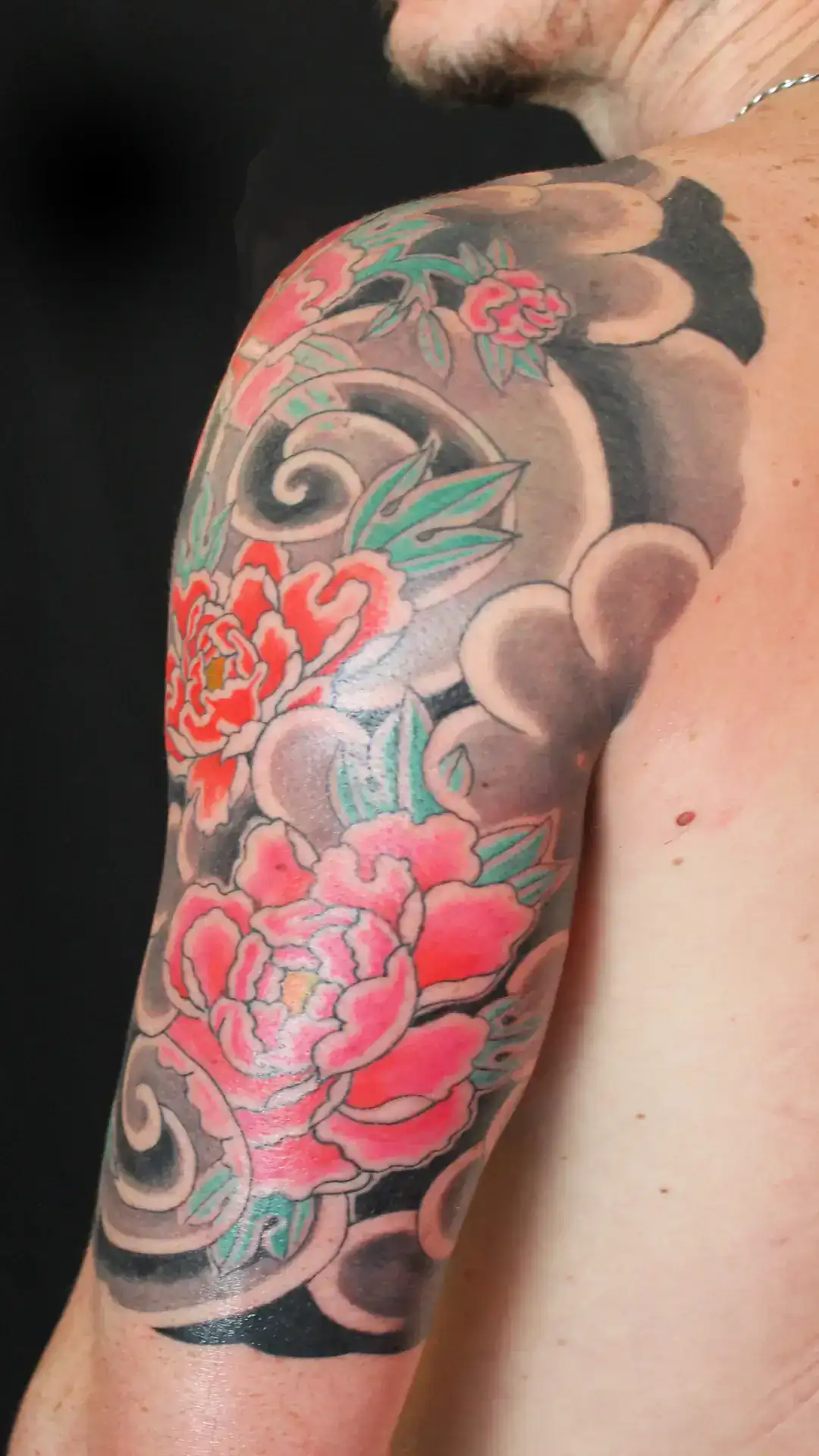 Detalle de tatuaje japonés de flores botan rojas con hojas verdes y nubes tradicionales en brazo, estilo irezumi por Nikko Quiroga.