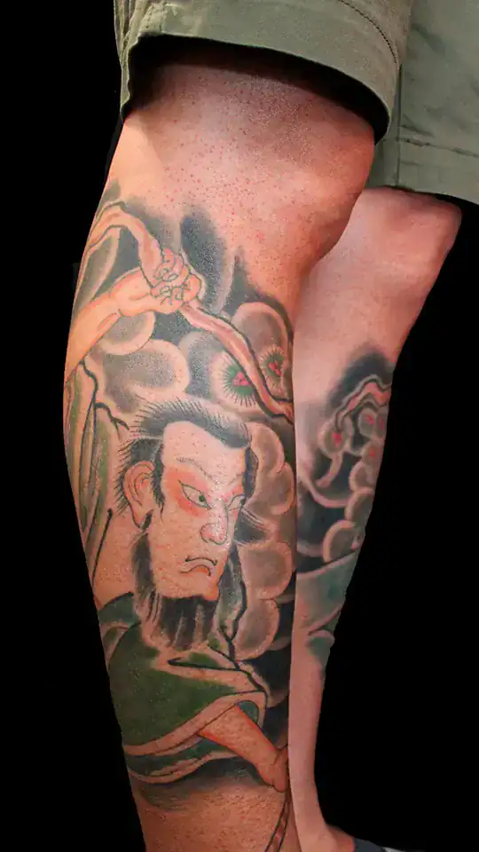 Primer plano del tatuaje de Musashi Miyamoto empuñando la espada, con fondo de nubes japonesas, en el gemelo, realizado por Nikko Quiroga.